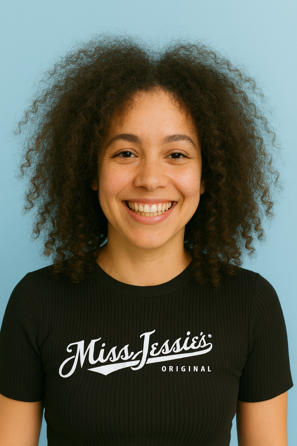 Smiling woman with natural curly hair wearing a black Miss Jessie’s Original shirt against a soft blue background, showcasing healthy, frizz-free curls for the blog How to Reduce Frizz for Natural Curls – Expert Tips